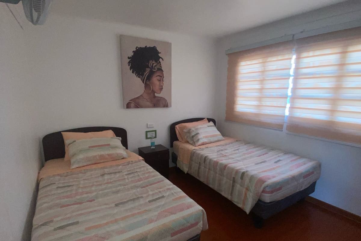 Un dormitorio con dos camas individuales, un ventilador de techo, una ventana con persianas y un cuadro colgado en la pared.