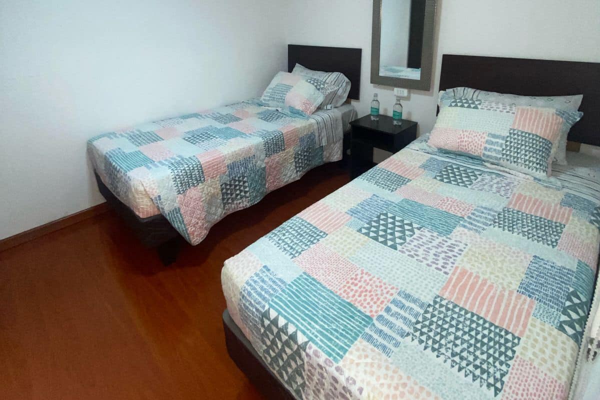 Habitación con dos camas, cada una con una colcha estampada, una mesita de noche con lámpara y un espejo montado en la pared.