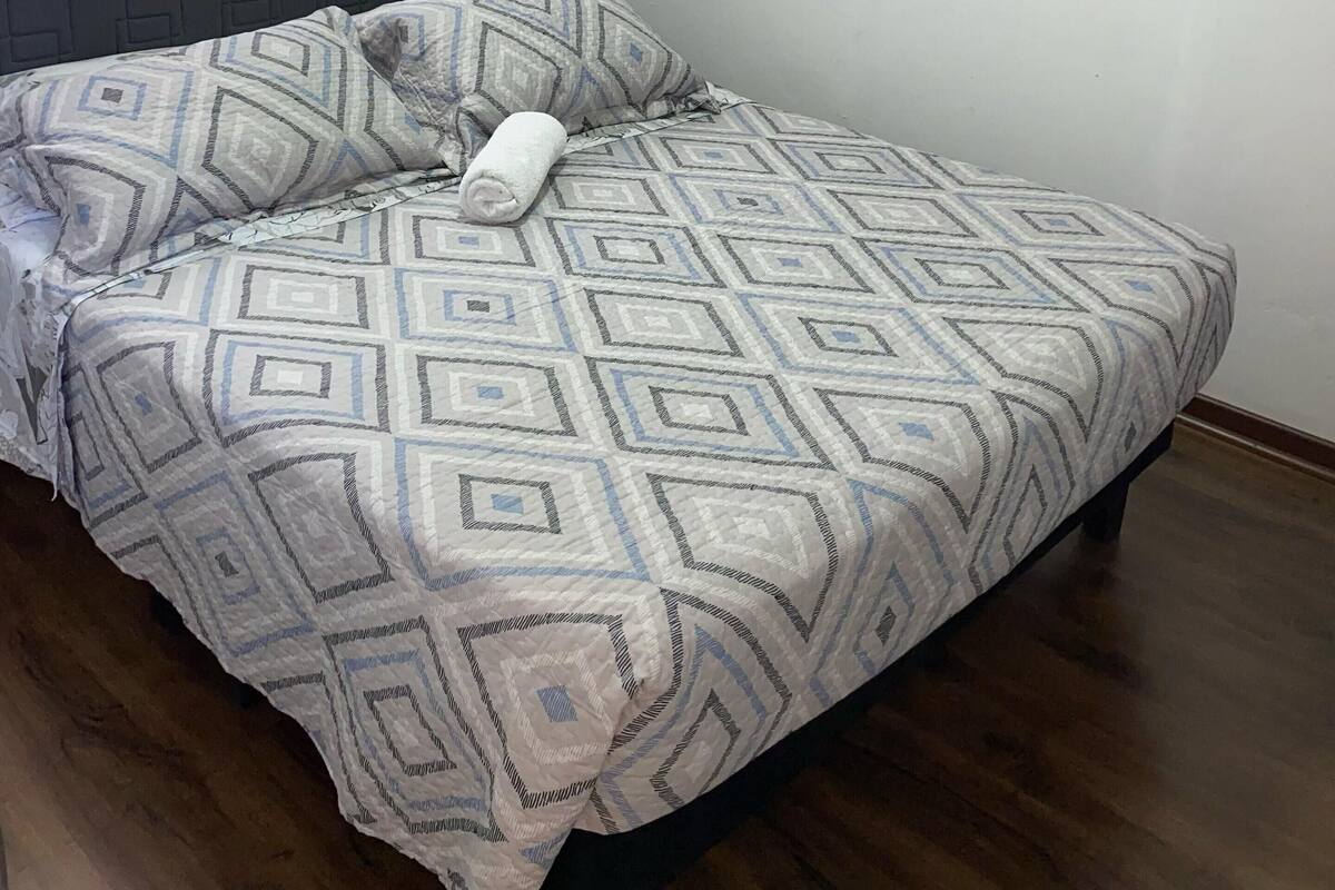 Un dormitorio con una colcha estampada, un cabecero, un cuadro en la pared y una ventana con persianas.