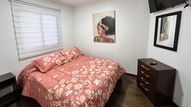 Un dormitorio con cama, cómoda, televisor montado en la pared y ventana con persianas.