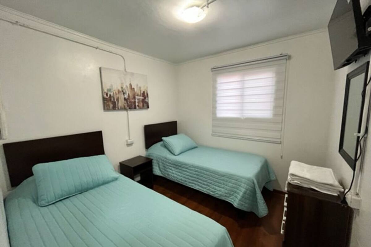 Una habitación de hotel con dos camas, una lámpara en el techo, una ventana con persianas, un cuadro en la pared y un televisor montado en la pared.