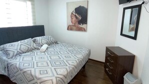 Un dormitorio con cama, cómoda, basurero, ventilador de techo, ventana con persianas y un cuadro en la pared.
