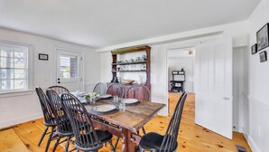 Un comedor con una mesa de madera dispuesta para cuatro personas, rodeada de sillas de metal. Se observa una zona de cocina con estantes que contienen vajilla y una estufa al fondo.