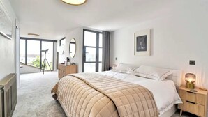 A modern bedroom with a large bed, a bedside table with a lamp, a dresser, and a large window offering a view of the outdoors. - Stunning House in Leigh-on-sea (Leigh-on-Sea)