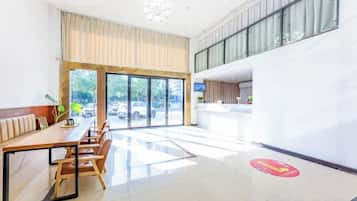 Um saguão de hotel com balcão de recepção, área de estar com uma mesa comprida e grandes janelas que oferecem vista para o exterior.
