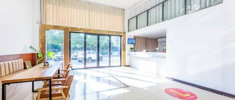 Un vestíbulo de hotel con una recepción, una zona de estar con una mesa larga y grandes ventanales que ofrecen vistas al exterior.