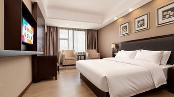 A hotel room with a large bed, a TV mounted on the wall, a desk with chairs, and a window with curtains.