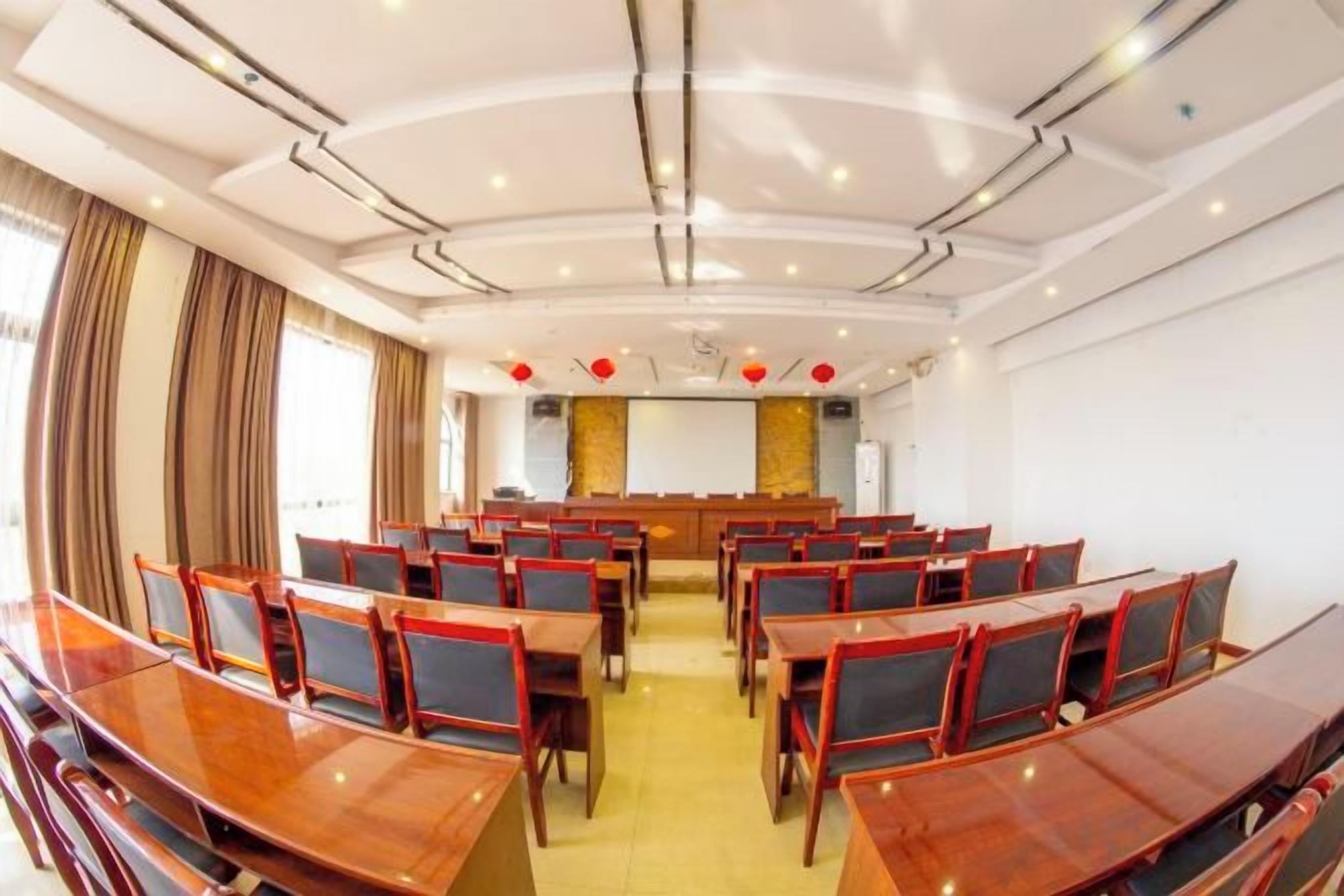 A conference room with rows of wooden tables and chairs, a stage with a podium, and a large screen at the front.