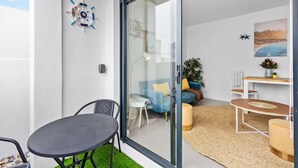 A modern living space with a glass door leading to a balcony, a blue sofa, a small table, and a potted plant. - Beach Break Two - One Bedroom Apartment, Sleeps 2 (Cape Town)
