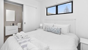 A modern bedroom with a large bed, a bedside table, a nightstand, a window, and a bathroom with a mirror and sink. - Beach Break Two - One Bedroom Apartment, Sleeps 2 (Cape Town)