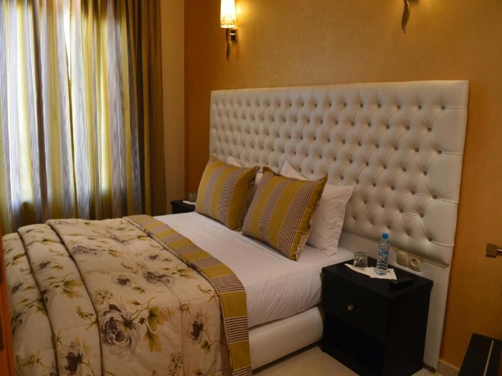 A hotel room with a large bed, a tufted headboard, a bedside table with a bottle and remote control, and a curtain on the window.