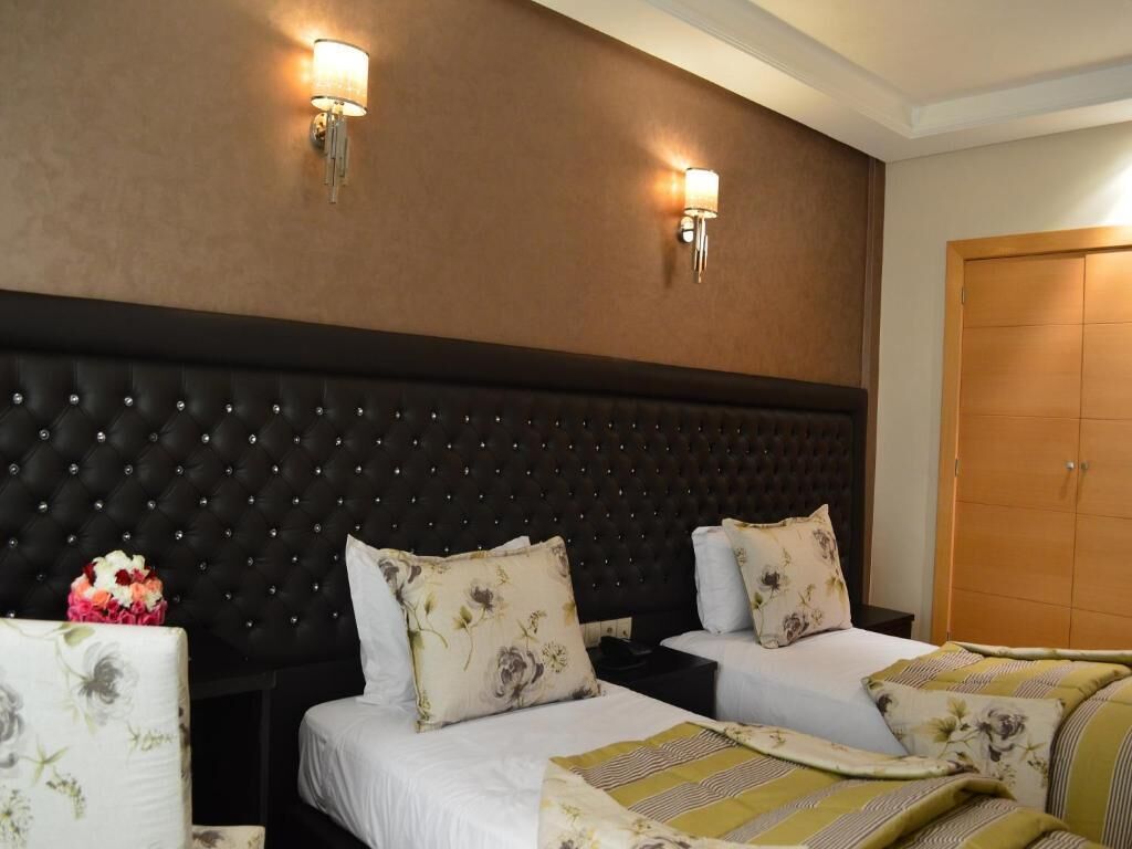A hotel room with a tufted headboard, two beds with floral bedding, a nightstand, and a wooden wardrobe.