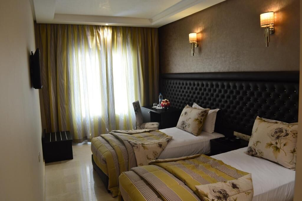 Habitación de hotel con dos camas, cabecera oscura, escritorio con silla y ventana grande con cortinas.