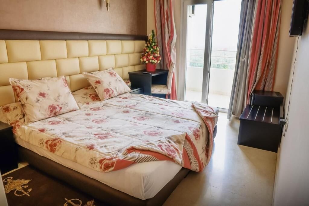 A hotel room with a large bed, a nightstand with a vase of flowers, and a view of the outdoors through a sliding door.