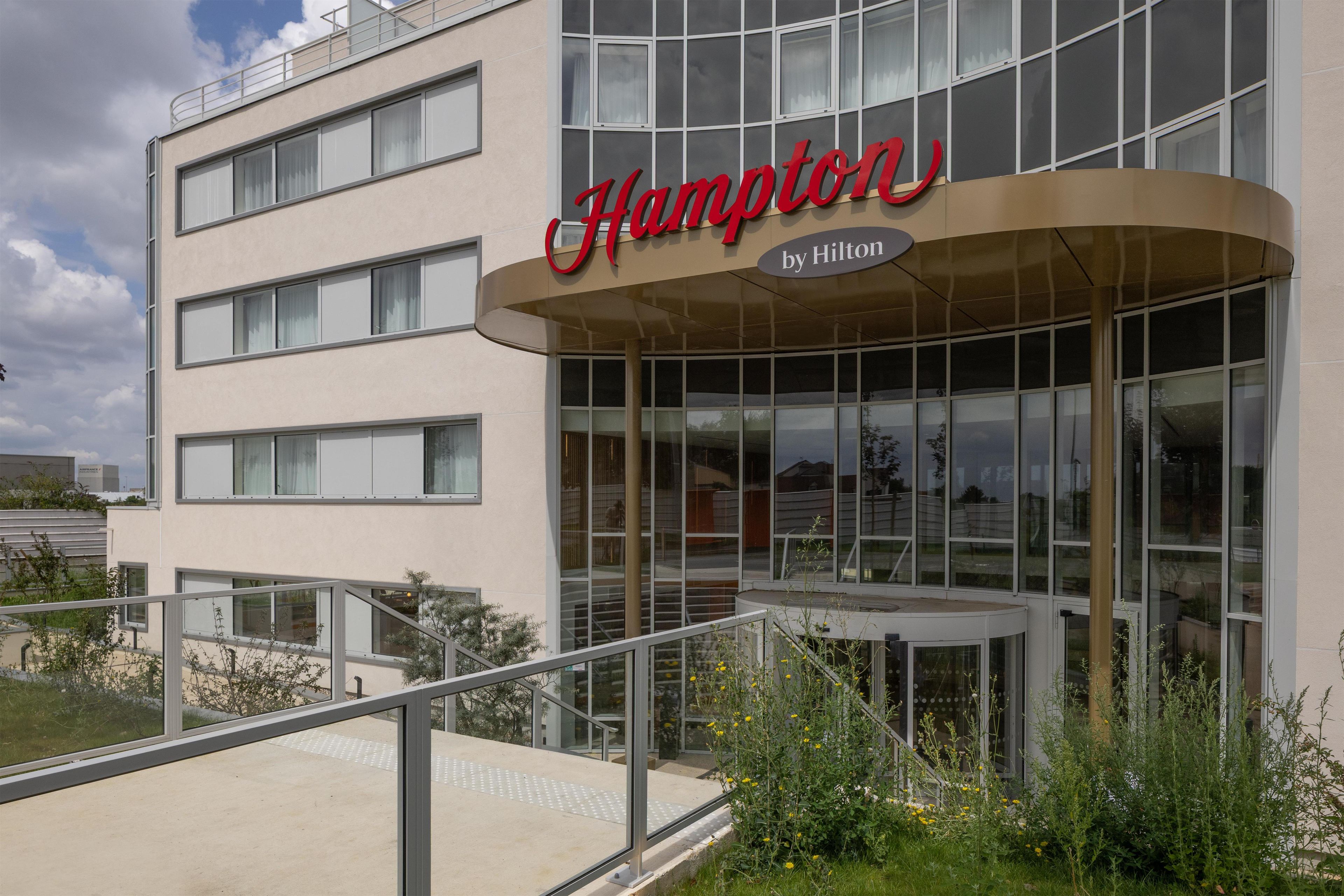 Um edifício moderno com um destaque para a placa 'Hampton by Hilton', grandes janelas e entrada de vidro.