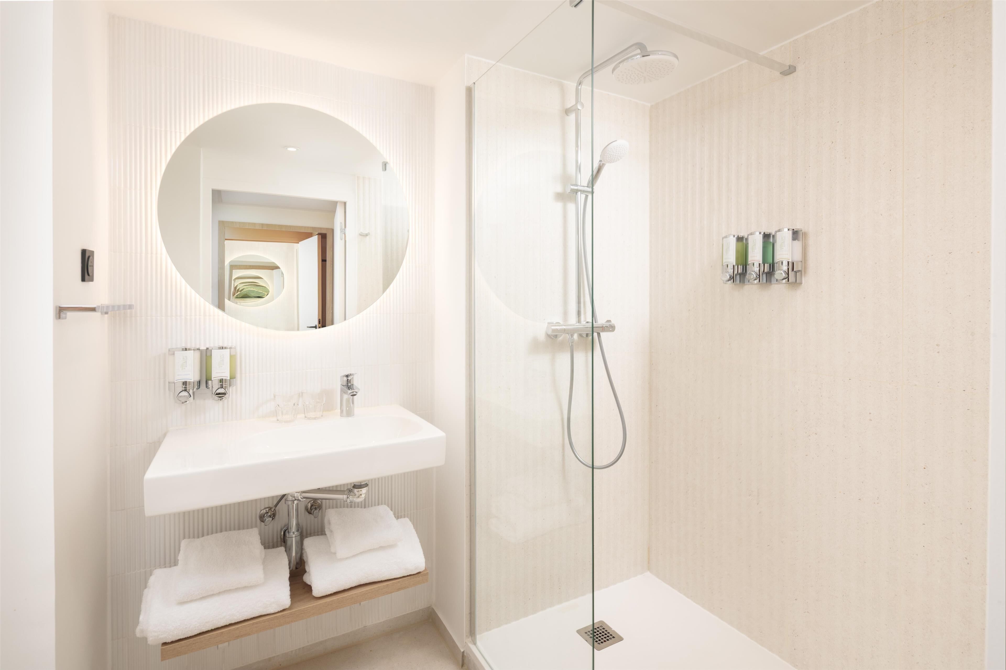 A modern bathroom with a glass-enclosed shower, a round mirror, a white sink, and a towel rack.