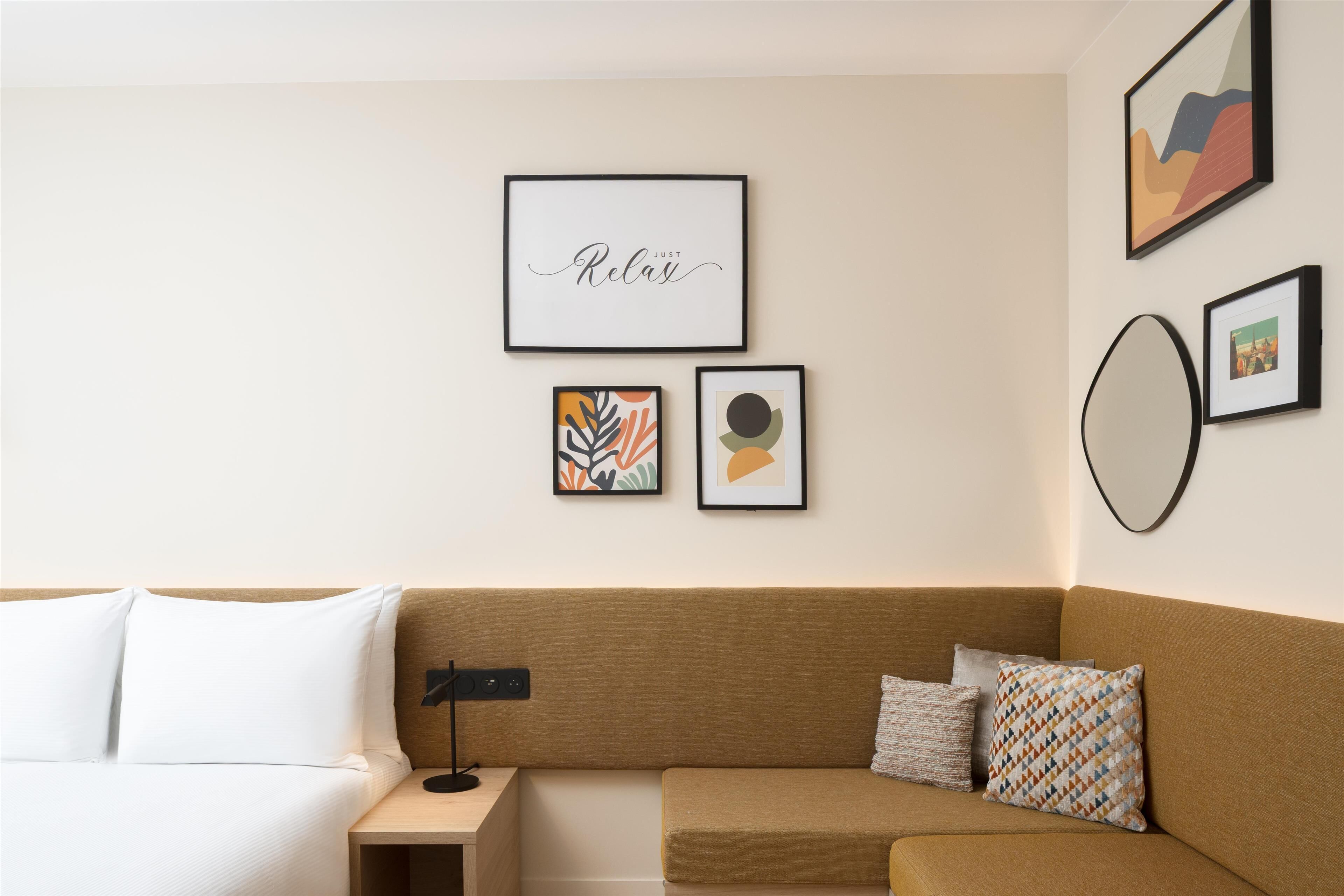 A modern hotel room with a brown sofa, a white bed, and a small nightstand. There are three framed artworks on the wall.