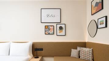 A modern hotel room with a brown sofa, a white bed, and a small nightstand. There are three framed artworks on the wall.