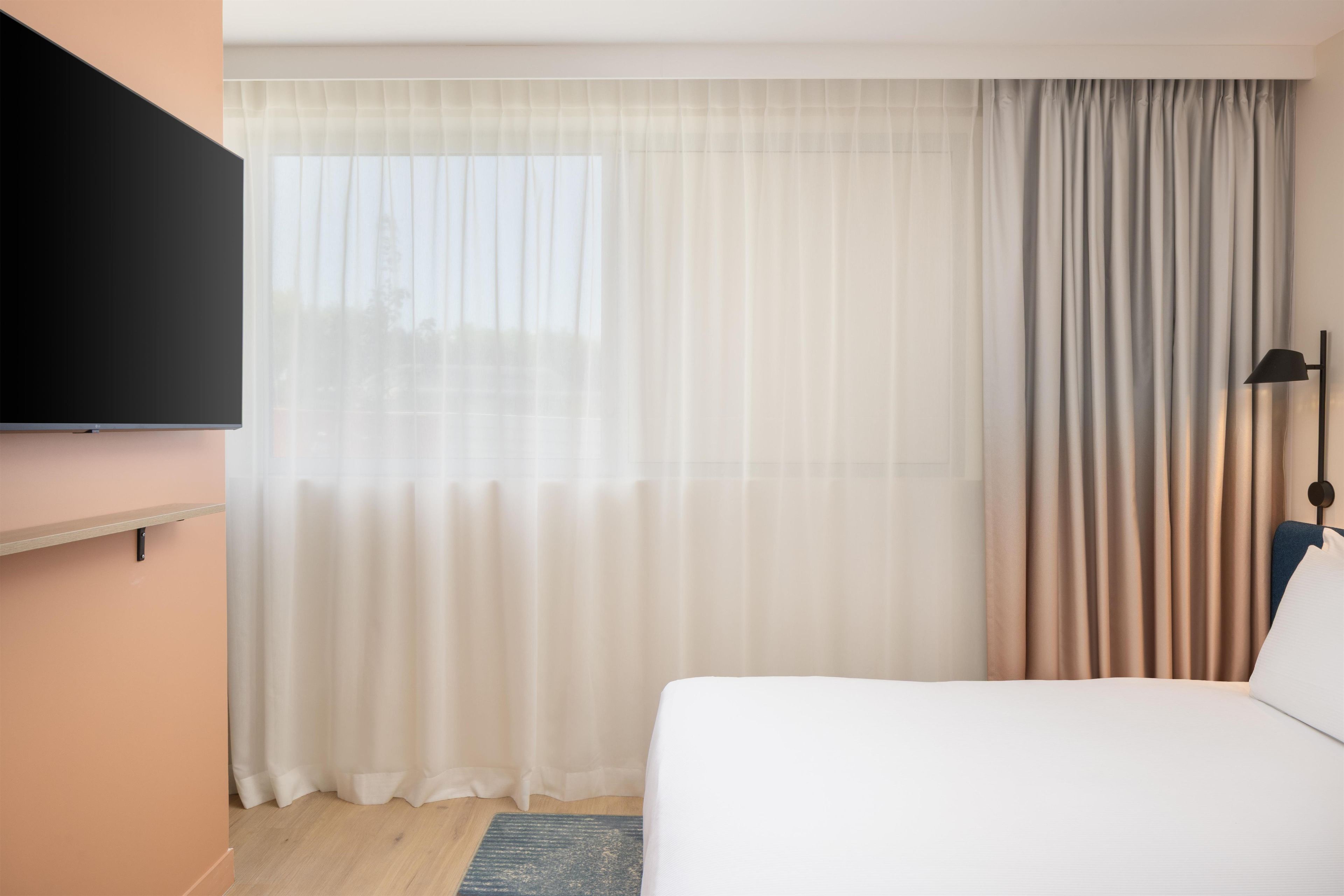 Una habitación de hotel con una cama, un televisor de pantalla plana montado en la pared, un ventanal con cortinas translúcidas y un juego de cortinas más oscuras.