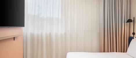 Una habitación de hotel con una cama, un televisor de pantalla plana montado en la pared, un ventanal con cortinas translúcidas y un juego de cortinas más oscuras.