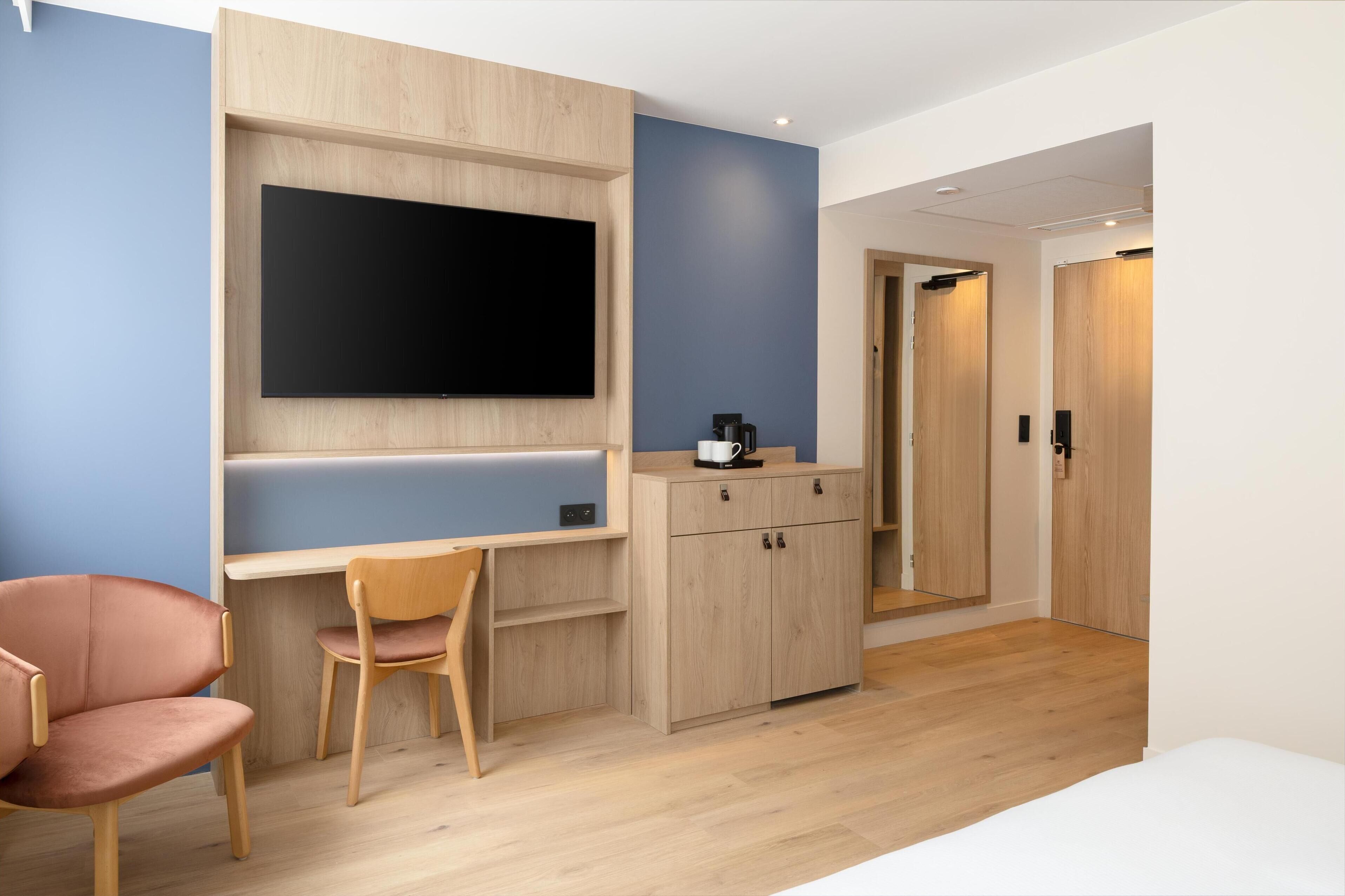 Quarto de hotel moderno com escrivaninha de madeira, TV de tela plana, máquina de café e armário.