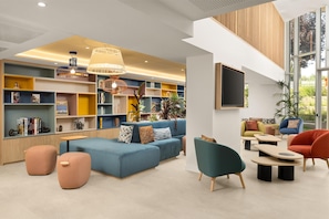 A modern lounge area with a blue sofa, teal armchairs, and a variety of bookshelves filled with books and decorative items. - Hampton By Hilton Paris Charles De Gaulle Airport (Roissy-en-France)