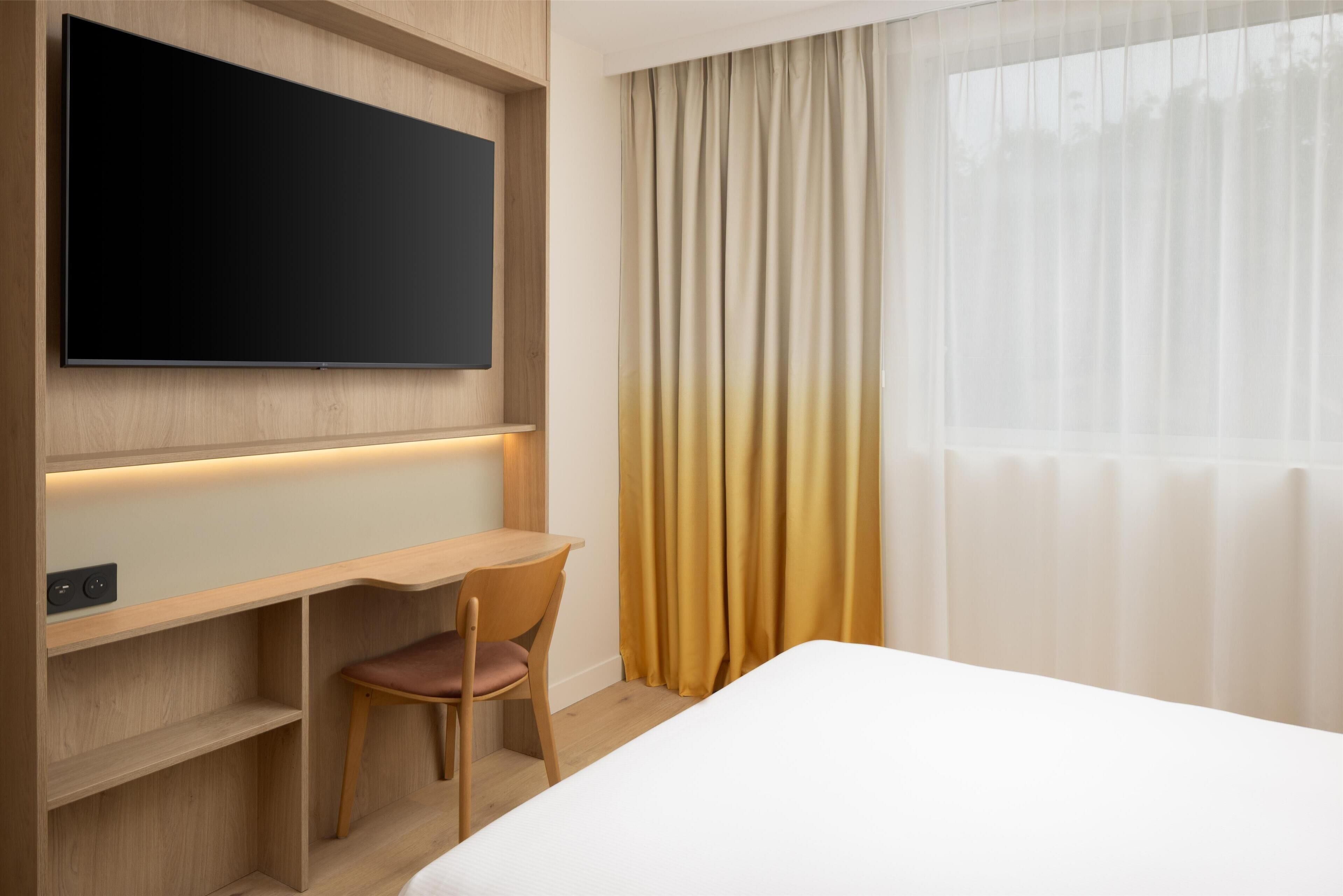 Habitación de hotel con un televisor de pantalla plana grande montado en la pared, un escritorio de madera con una silla y una cama con colchón blanco.