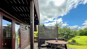 Une terrasse en bois avec une table et des bancs, une porte-fenêtre coulissante et une vue sur un paysage verdoyant.