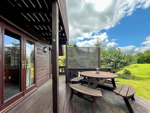 Een houten terras met een tafel, banken, een schuifpui en uitzicht op een groen landschap.