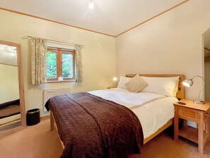 A bedroom with a bed, bedside table, lamp, window with curtains, radiator, and a mirror.