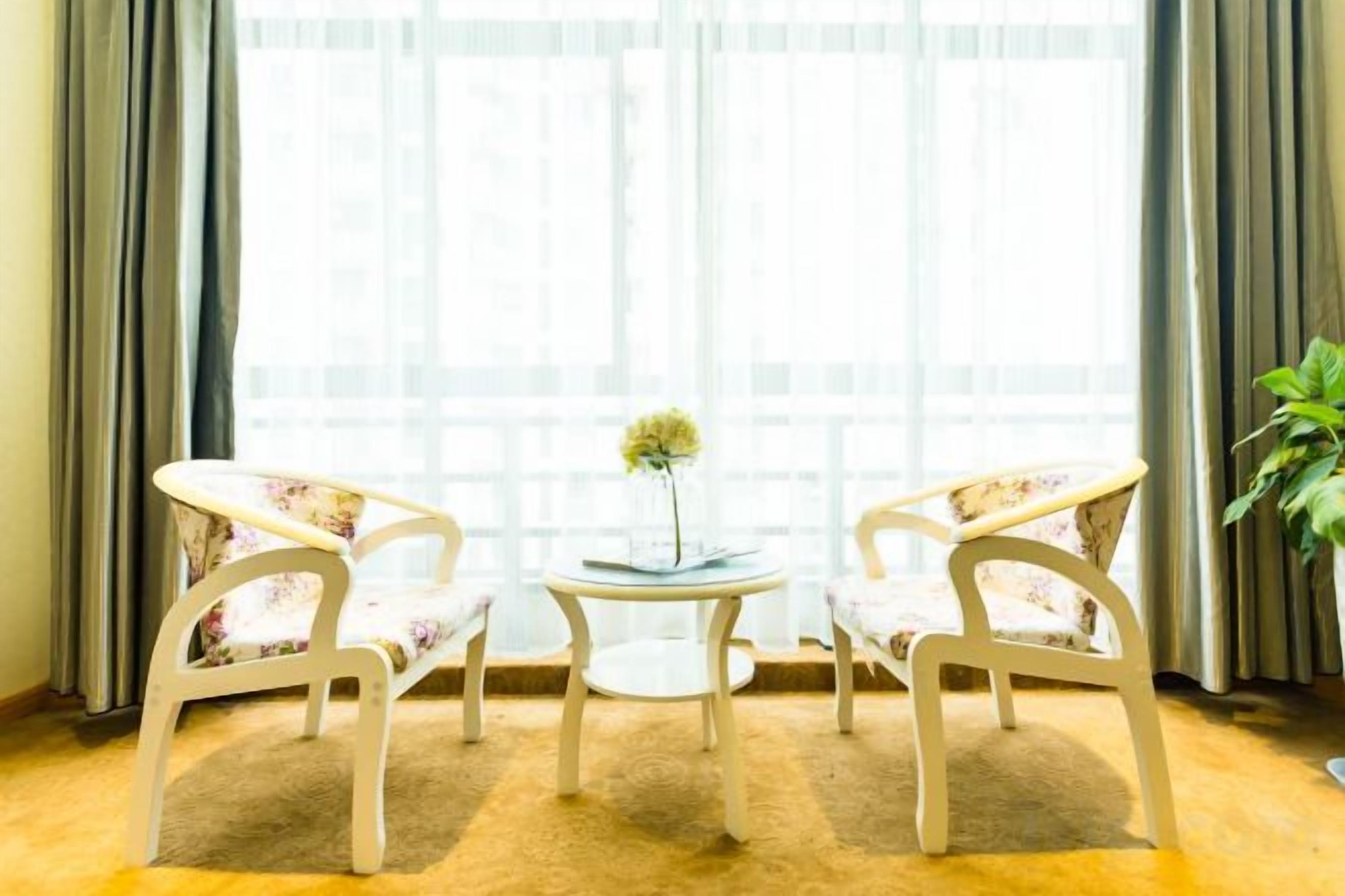 Une pièce avec deux chaises blanches, une petite table ronde surmontée d’un vase et une grande fenêtre avec des rideaux.