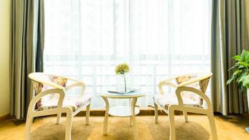 A room with two white chairs with floral upholstery, a small round table with a vase of flowers, and a large window with curtains.