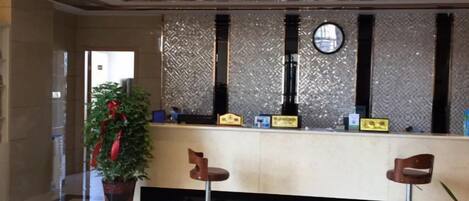 Zona de recepción con mostrador de check-in, dos taburetes de bar, una planta en maceta y una pared decorativa con un reloj.
