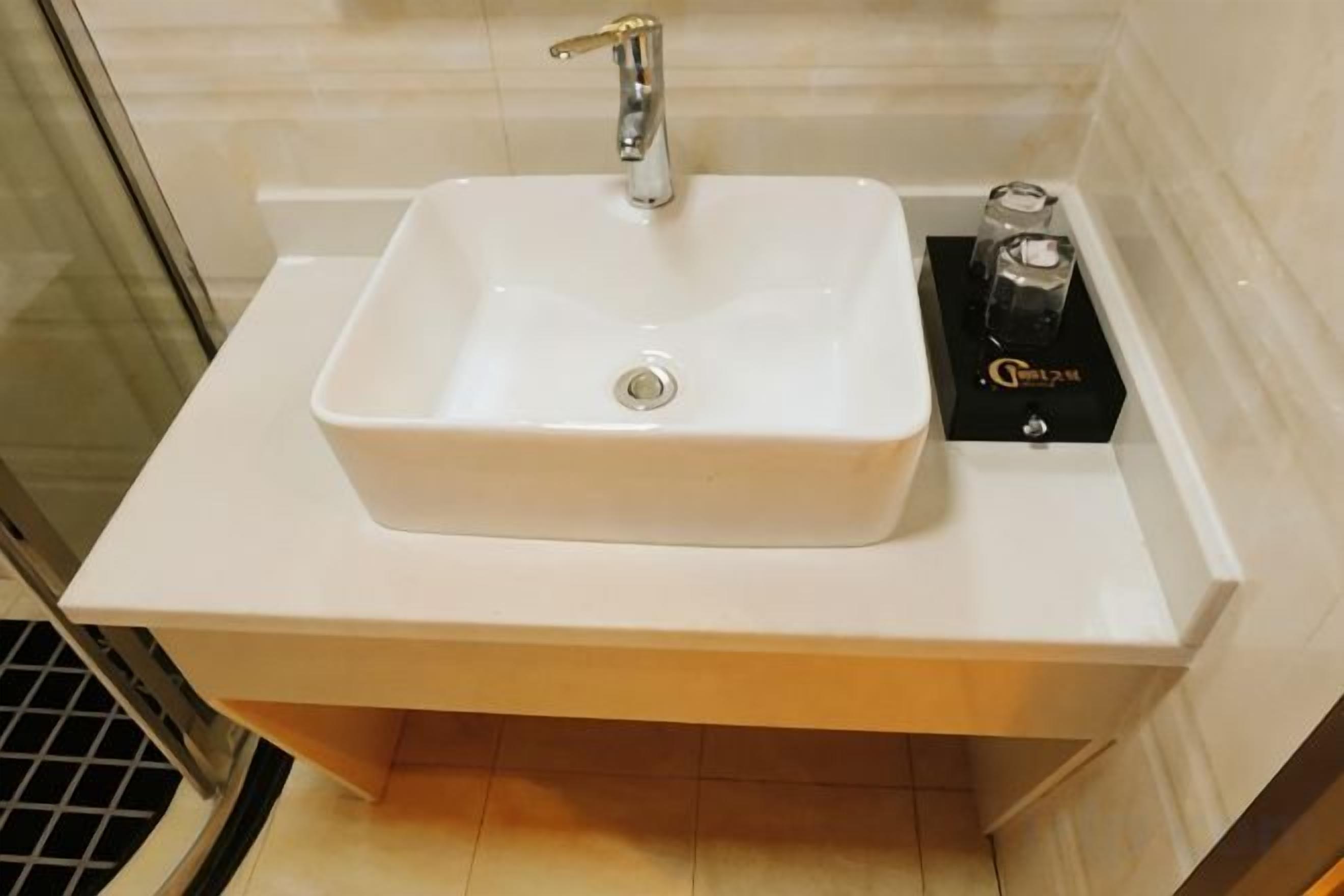 A white rectangular sink with a single faucet, a black box with gold lettering, and a tiled wall in the background.