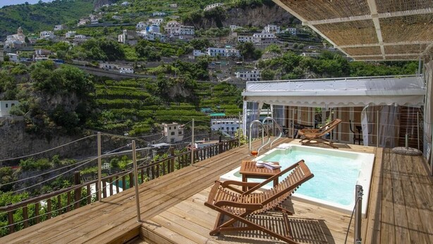 Une terrasse en bois avec une piscine, des chaises longues et une vue sur un village perché sur une colline.