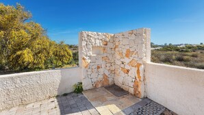 Estructura de pared de piedra con una pequeña abertura, rodeada por una zona pavimentada y una pared blanca.