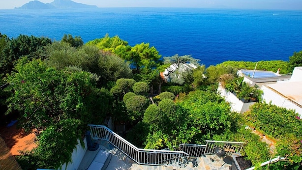 Une propriété située sur la côte, dotée d’un escalier menant à un espace sur le toit, entourée d’une végétation luxuriante et offrant une vue sur la mer et les montagnes lointaines.