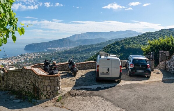 A stone wall with parked motorcycles and cars, overlooking a coastal town and mountains.