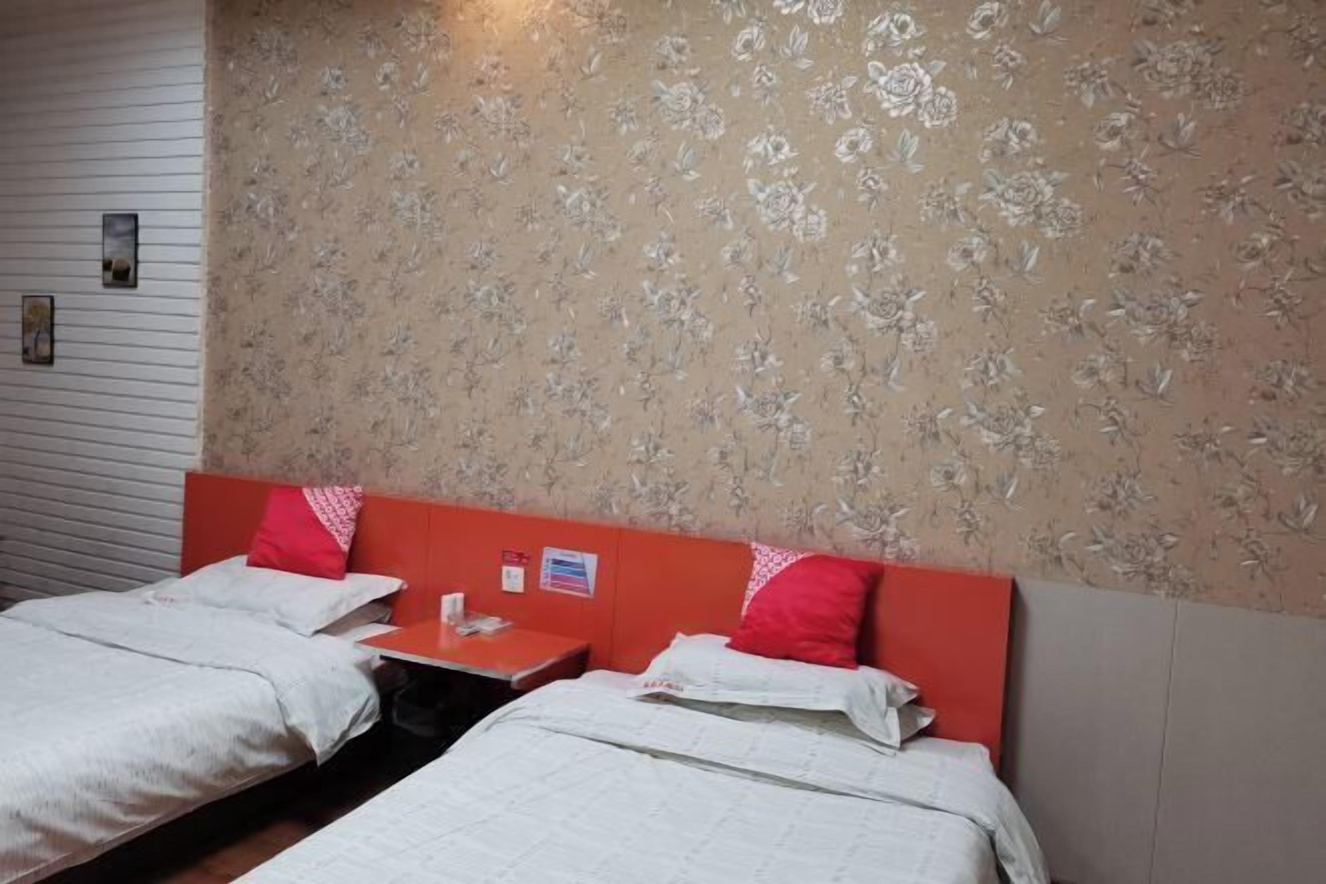 Habitación de hotel con dos camas individuales, cabecera roja, papel tapiz con motivos florales y una pequeña mesa con cojín rojo.