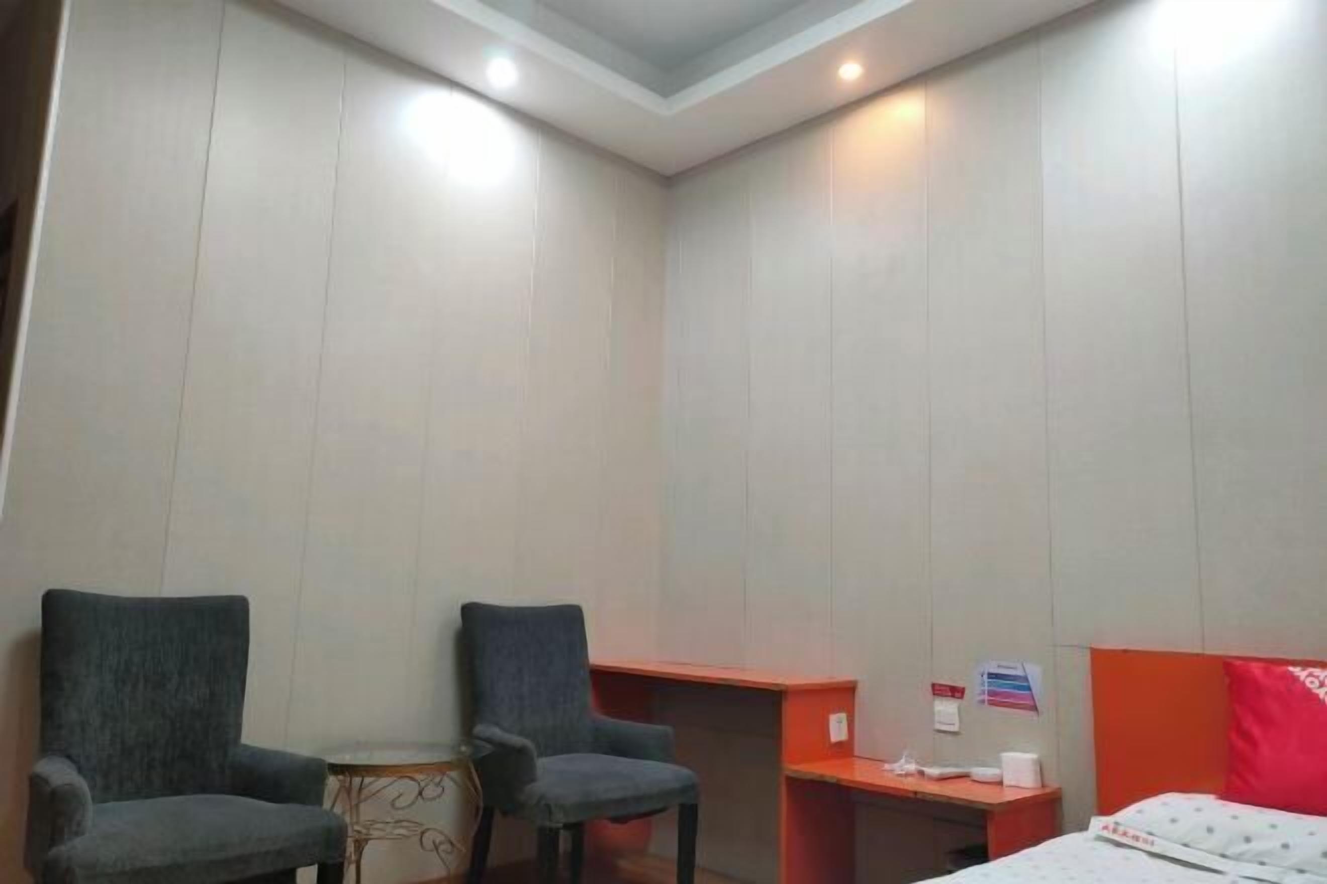 A hotel room with two armchairs, a small table, a desk, a bed with a red headboard, and a bulletin board.