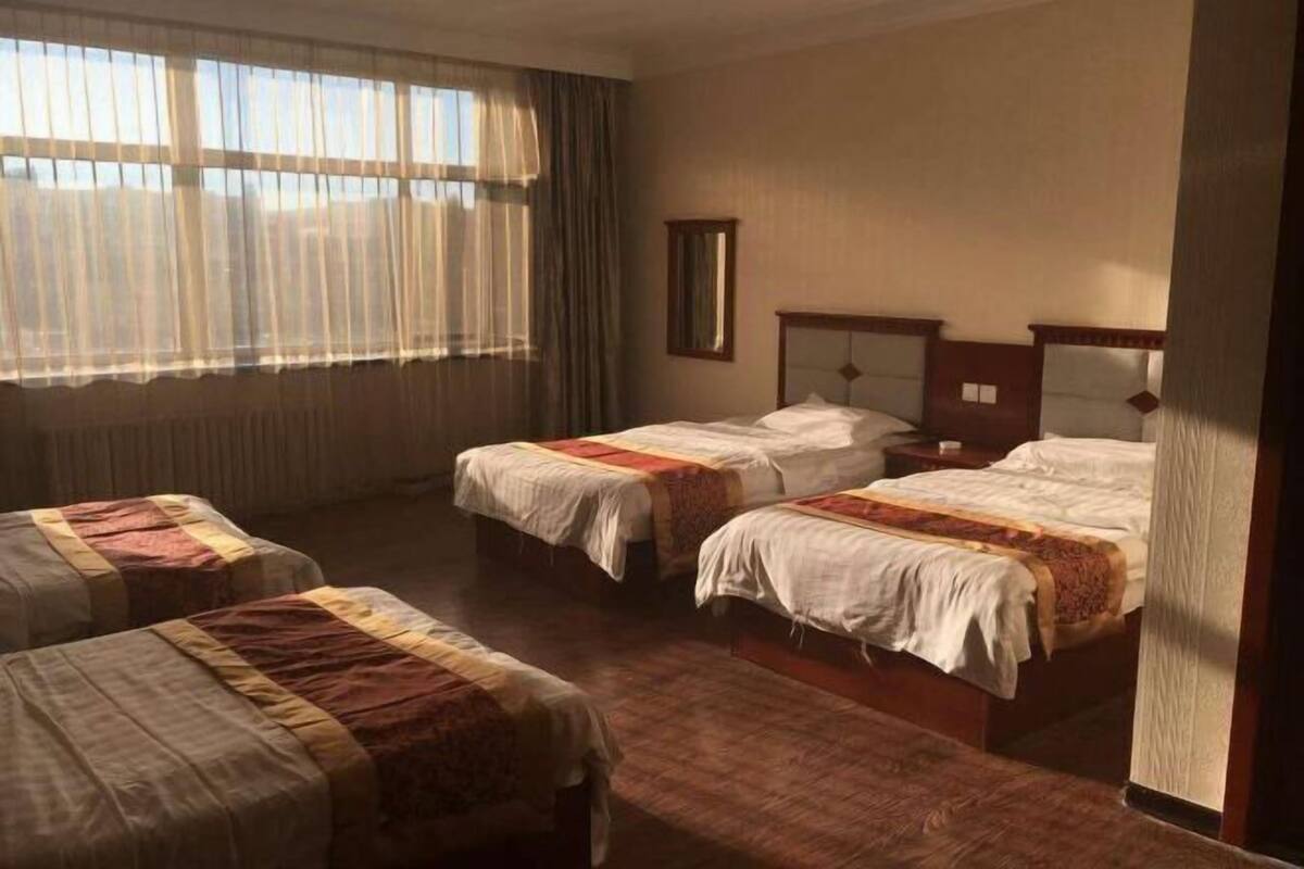 Habitación de hotel con dos camas individuales y una cama doble, un ventanal con cortinas translúcidas y suelo de madera.