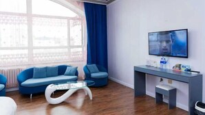 A modern living room with a blue sofa, a glass coffee table, a TV on a stand, and a window with curtains.