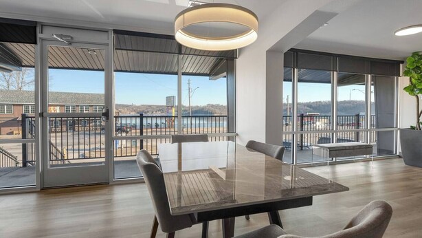 Ein moderner Essbereich mit Glastisch und Stühlen, großen Fenstern mit Blick auf ein Gewässer und Gebäude, sowie einer hängenden Leuchte.