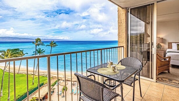 A balcony with a view of the ocean, a dining table with chairs, and a bedroom with a bed and a nightstand.