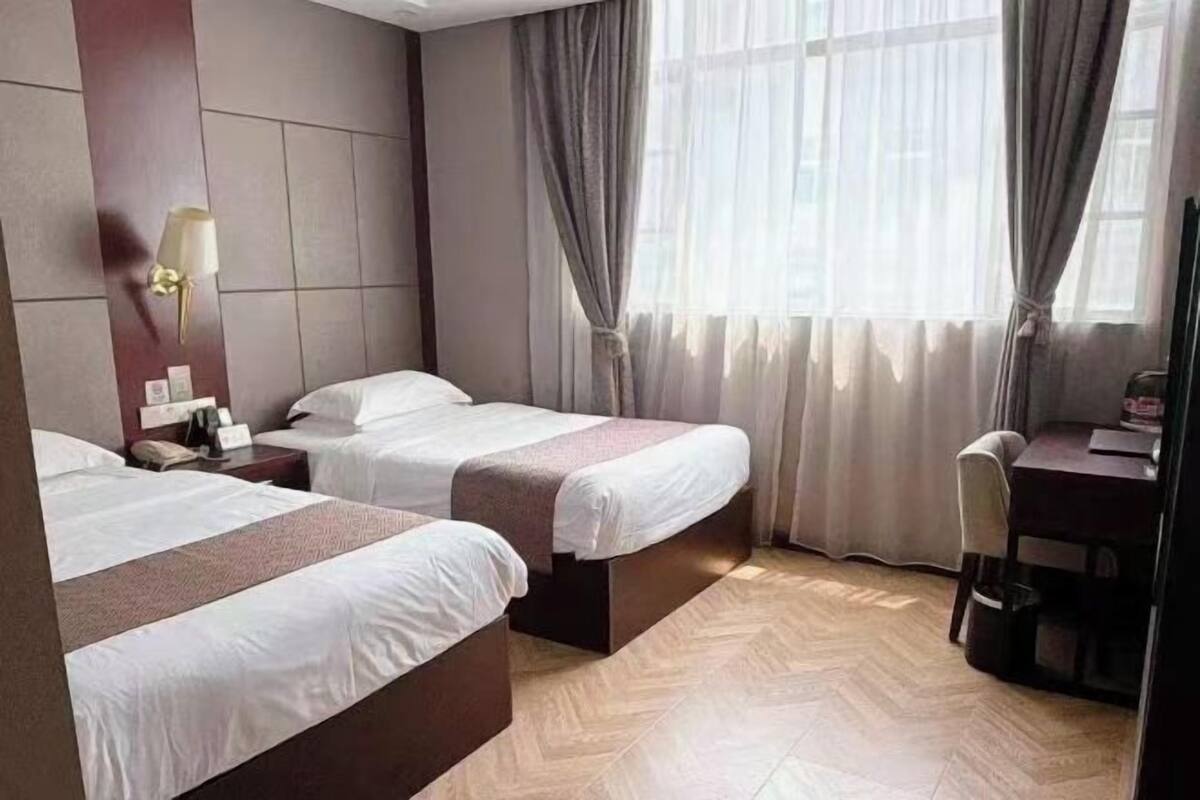 Habitación de hotel con dos camas, un escritorio, una silla y un amplio ventanal con cortinas.