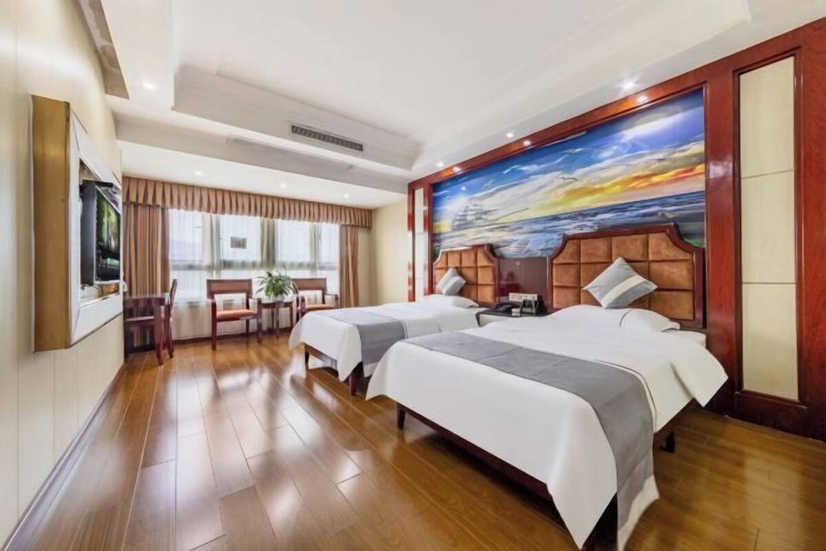 Habitación de hotel con dos camas, un cuadro grande de un paisaje marino, suelo de madera y una zona de comedor con una mesa y sillas.