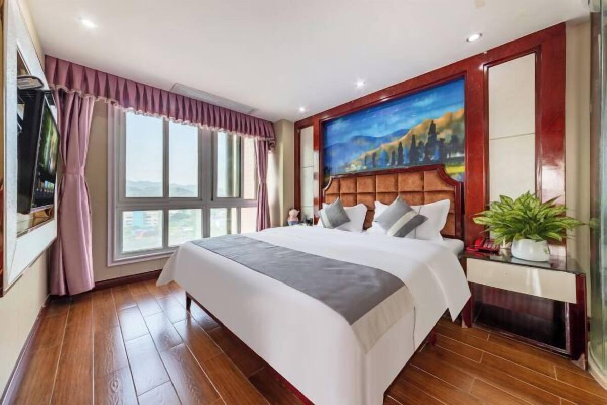 Una habitación de hotel con una cama grande, un cuadro en la pared, una ventana con cortinas y una planta sobre una mesita.