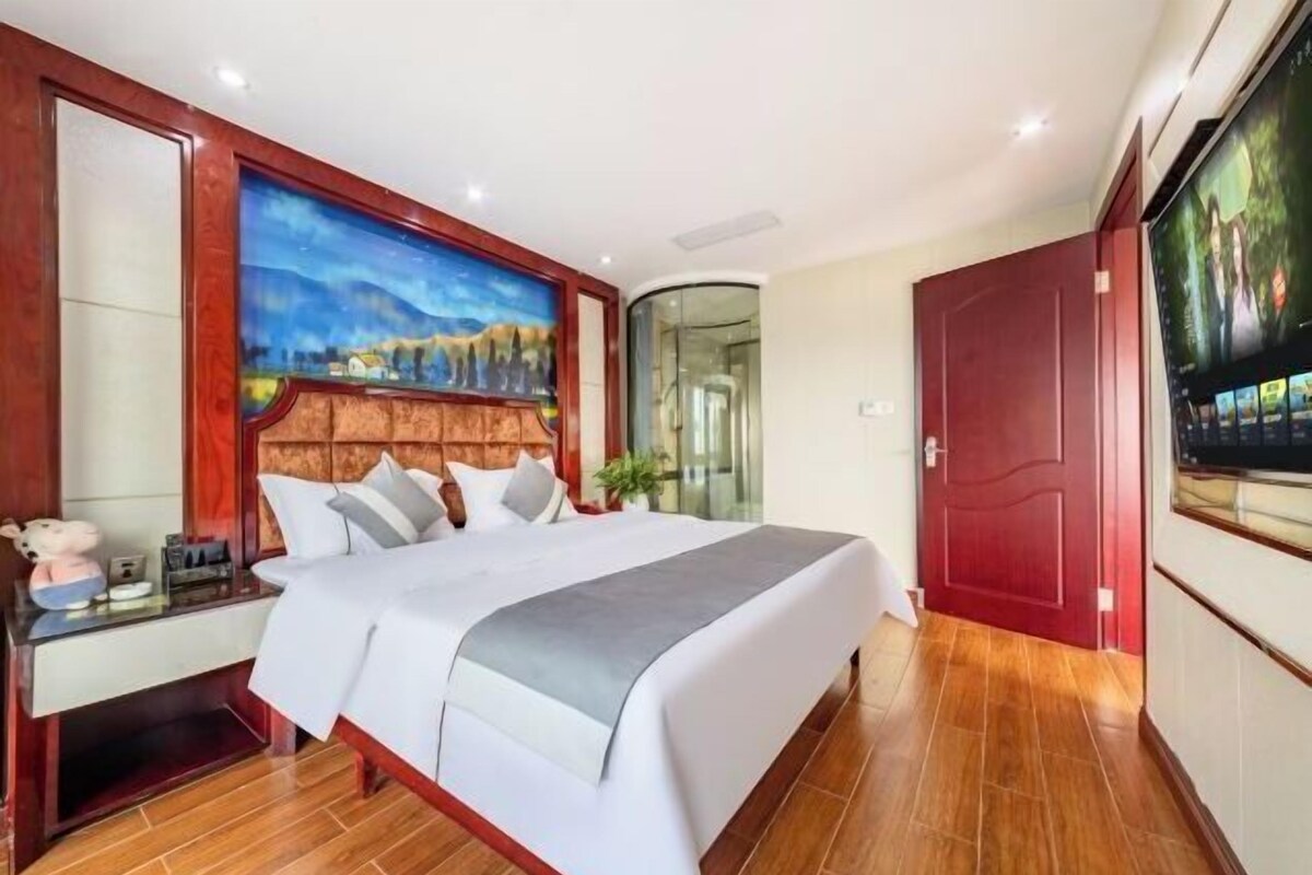 A hotel room with a large bed, a desk with a chair, a painting on the wall, and a TV on the wall.
