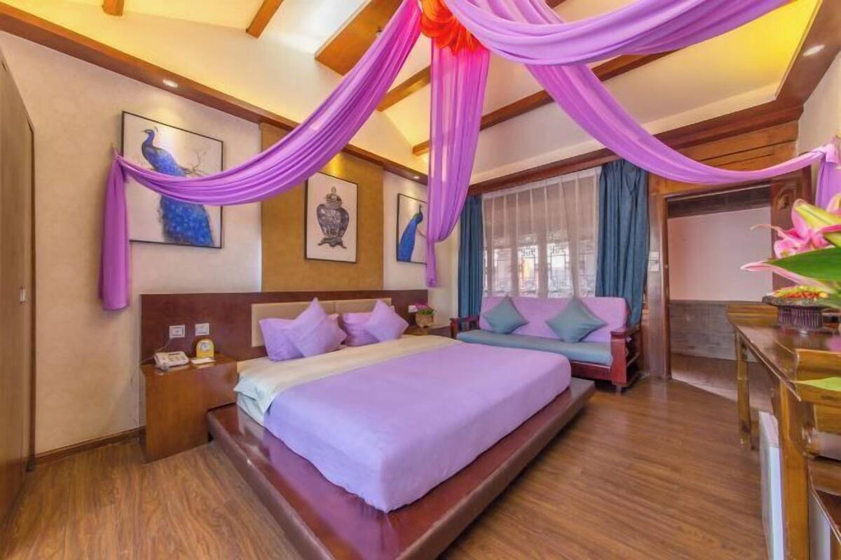 A hotel room with a large bed, wooden headboard, and purple drapes. There are framed pictures on the walls and a window with curtains.