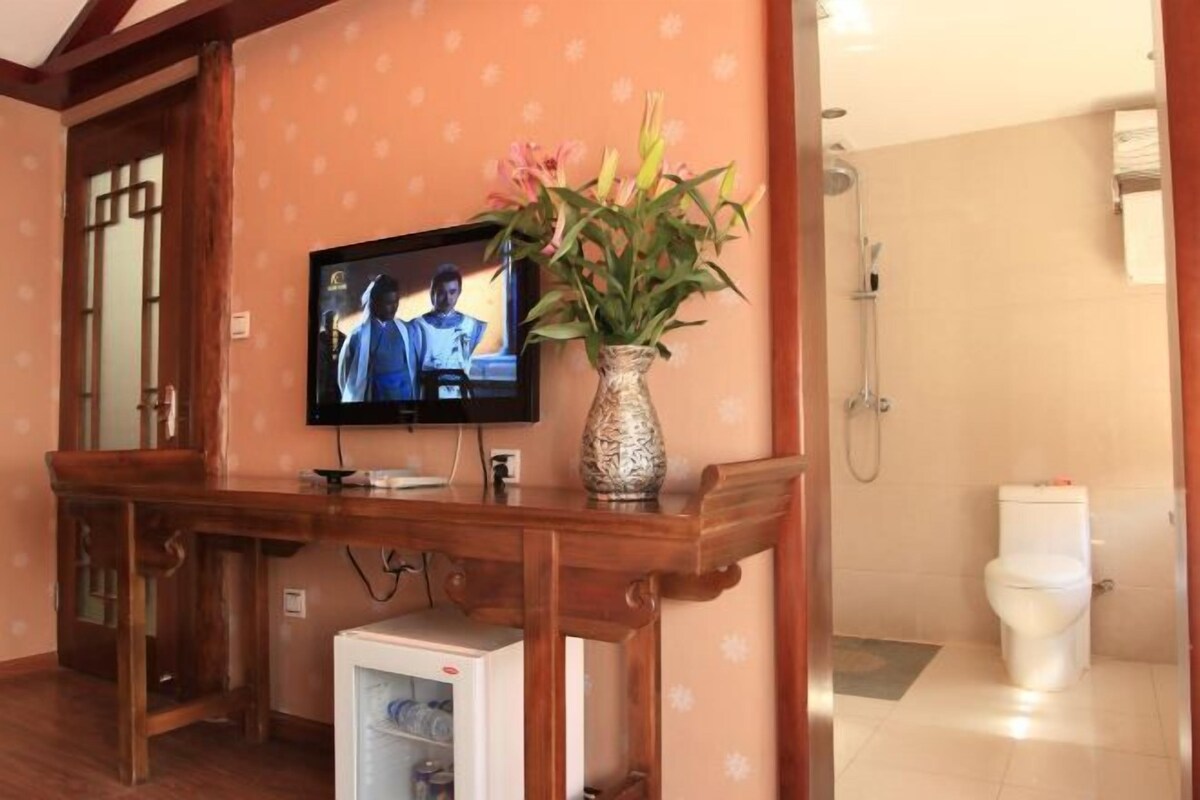 Quarto com escrivaninha de madeira, televisão, vaso de flores, pequeno frigorífico e sanita.